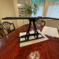 A black adjustable laptop stand placed on a polished wooden dining table in a bright dining room with windows and a china cabinet