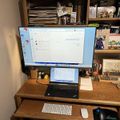 A home office workspace with a laptop connected to a large external monitor, wireless keyboard and mouse on a wooden desk, with bookshelves containing papers and books in the background