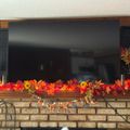 A fireplace mantel decorated with colorful autumn leaves and flowers, with a large TV mounted above it and decorative lanterns on either side