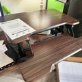 A height-adjustable standing desk converter with wood-grain finish positioned on top of a regular desk, with instruction manual and office supplies visible in the workspace