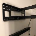 Black TV wall mount bracket installed on white wall with soundbar mounted below and visible cables