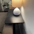 A white ceramic table lamp with textured base and beige lampshade sits on a dark wooden side table next to a wall, with plants visible in the background