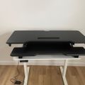 A black adjustable standing desk converter with two tiers positioned on a white desk frame, with a power cord visible on the wooden floor