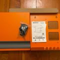 Orange wall mount shelf kit with installation hardware and instruction manual laid out on wooden floor