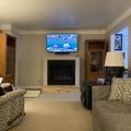 A cozy living room with a fireplace, mounted TV showing a weather forecast, comfortable seating including a patterned chair and gray sofa, wooden built-in cabinets, and warm lighting from a table lamp.