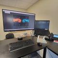 Office workspace with dual monitor setup showing colorful graphics on large wall-mounted display, laptop, keyboard, mouse, and phone on dark desk