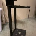 A black metal stand or frame with an open rectangular structure, featuring a hinged top panel and a base platform with geometric cutout patterns, positioned on a carpeted floor against a beige wall.