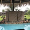 Backyard pool area with tiki-style thatched roof pavilion, stone edging, and tropical landscaping including palm trees