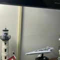 A display shelf showing LEGO models including a lighthouse with red roof and a Star Wars Imperial Star Destroyer on a black stand, with a TV visible in the background