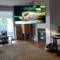 Living room with large wall-mounted TV above fireplace, wooden floors, woven area rug, dining table, and moving boxes stacked nearby