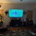 Living room with large TV displaying a football game, featuring a coffee table, patterned rug, ceiling fan, and various furniture and decor items