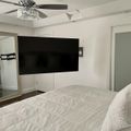 Modern bedroom with white bedding, wall-mounted TV, ceiling fan with crystal light fixture, and white walls with decorative molding