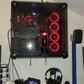 A gaming setup with a wall-mounted PC case featuring red LED fans, a desk with headphones, phone, and gaming accessories in a white room