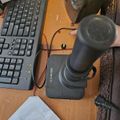 A desktop setup viewed from above showing a black keyboard on the left, a wooden desk surface, a joystick controller in the center, computer speakers in the background, and various desk accessories including a white paper and fabric items on the right side.