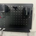 Black metal pegboard organizer with various tool holders and accessories mounted on a stand, positioned on a checkered surface against a white wall