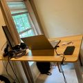 A wooden desk with a laptop, cables, and phone positioned near a window with curtains in a home office setting