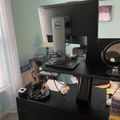 A home office workspace with a standing desk setup featuring a Dell monitor, laptop, desk lamp, and various office supplies on a black desk surface near a window with blinds.