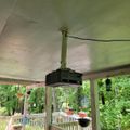 A projector mounted on a ceiling mount under a covered porch or deck area with white railings and green foliage visible in the background