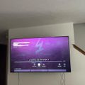 A large flat-screen TV mounted on a white wall displaying a purple-themed media interface with playback controls and song information