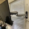 A computer monitor mounted on an adjustable arm next to a window with white horizontal blinds in what appears to be a home office or workspace