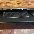 A black keyboard tray pulled out from under a wooden desk with a wireless keyboard and mouse on top