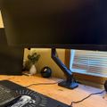 A computer workstation with a large monitor on an adjustable arm, wooden desk surface, laptop with spiral design on screen, white mouse, small decorative vase with plant, and window with blinds in background