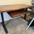 A wooden desk with black metal legs and a pull-out keyboard tray, positioned on beige carpet in a room with storage boxes visible in the background.