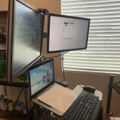A multi-monitor computer workstation setup with three screens mounted on adjustable arms, a keyboard, and desk accessories positioned near a window with white blinds