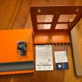 Unboxed orange and brown wall mount bracket with installation hardware, instruction manual, and mounting screws laid out on wooden floor
