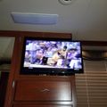 Interior of an RV or motorhome showing a wall-mounted TV displaying a football game, with wooden cabinetry and window blinds visible