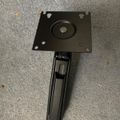A black metal monitor stand or mount with a square base plate and adjustable arm lying on a gray carpeted surface