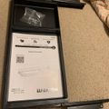 An open black box containing assembly instructions and a plastic bag with hardware components, sitting on beige carpet