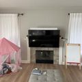 Living room with fireplace, mounted TV, white curtains, pink play tent, easel, and gray ottoman on hardwood floors