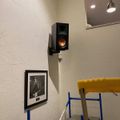 Interior room with white textured walls featuring a wall-mounted black speaker, framed black and white photograph, yellow chair, and blue metal frame structure