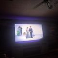 A projected image on a wall showing an animated couple in formal attire standing together in what appears to be a wedding scene, displayed in a dimly lit room with purple ambient lighting