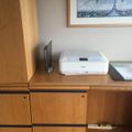 A wooden desk or cabinet with a white Canon printer on top, alongside some papers and office supplies, with a framed Paris cityscape artwork hanging on the white wall above