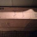 A cardboard box for a Mount-It L-shaped sit stand desk, model MI-7902, sitting on what appears to be a tiled floor against a ceiling