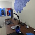 A modern office workspace with a curved wooden desk, computer monitor on an adjustable arm, laptop, blue ceramic pen holder, and decorative blue geometric wall panels