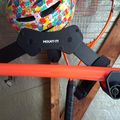 A colorful puzzle-patterned helmet sits on an orange bike frame with black mount components, positioned near wooden furniture on a tiled floor.