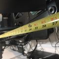 A measuring tape extended to show measurements around 24-27 inches, positioned near what appears to be electronic equipment or machinery in a workspace setting.