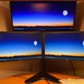 Three computer monitors displaying a scenic desktop wallpaper of a sunset/sunrise landscape with mountains, blue sky, and full moon, arranged in a multi-monitor setup on a wooden desk