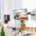 A person sitting on a couch watching a wall-mounted TV displaying a gray cat being petted by a human hand in a modern living room with plants and furniture
