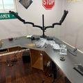 A workspace desk with computer equipment including a dual monitor arm mount, various cables, small toy vehicles, and packaged items. Street signs including "DAMEN AV" and a STOP sign are visible on the wall above the desk.