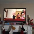 TV on white fireplace mantel showing animated movie with baby character, Christmas stockings hanging below