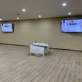 Empty room with beige walls, wood-look flooring, recessed lighting, two wall-mounted TV screens, and a wrapped piece of equipment or furniture in the center
