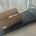 A cardboard box sitting on top of a black treadmill near a window with white curtains on a wooden floor