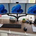 A home office desk setup with three monitors displaying blue abstract wallpapers, a keyboard, mouse, phone, and various office supplies on a wooden desk surface