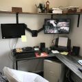 A home office workspace with dual monitors on an adjustable arm mount, an L-shaped desk with keyboard and office supplies, a white office chair, and a wooden shelf above containing various items including books and a mug.