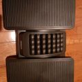 Three black kitchen appliance plates or grilling surfaces stacked on a wooden floor, with the middle one showing a waffle-making pattern of round indentations