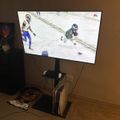 A television mounted on a stand displaying an NFL football game with players on a snowy field, with gaming consoles visible below the TV